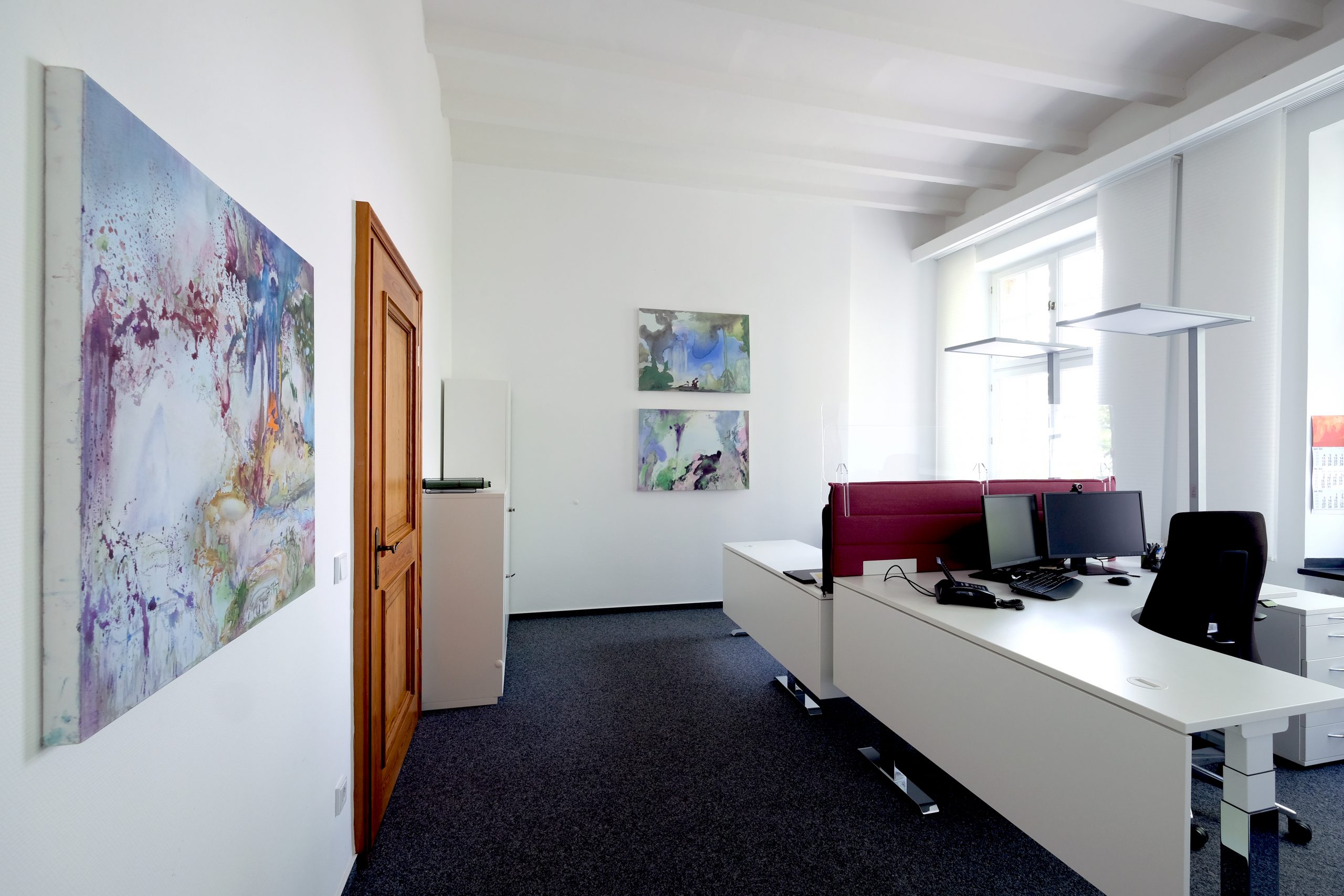 ein Büroraum mit Schreibtischen, Bildschirmen und drei Bildern an der Wand