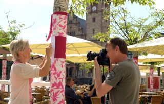 Eine Frau bringt ein Stoffband an einen Baum an und ein Mann fotografiert sie dabei.