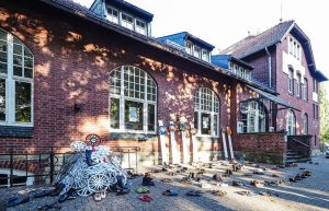 Fotografie einer Installation vor einem Bahnhofsgebäude. Man sieht einen Hügel aus Radkappen sowie fein säuberlich hintereinander aufgereihte Paar Schuhe.