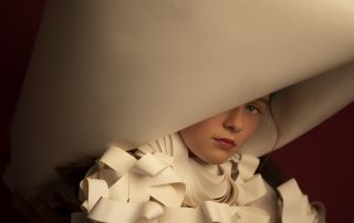 Der Hut und die Kleidung eines jungen Mädchens besteht aus Papier. Sie schaut in die Kamera, aber der Hut verdeckt ihr rechtes Auge.