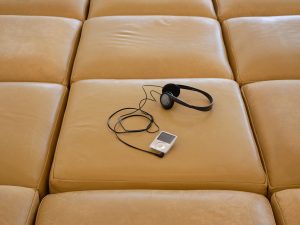 ID: Ein an einen iPod angeschlossener Kopfhörer liegt auf einer gitterartigen Anordnung von großen, quadratischen Lederkissen in hellbrauner Farbe. Die Kissen weisen sichtbare Falten und Gebrauchsspuren auf.